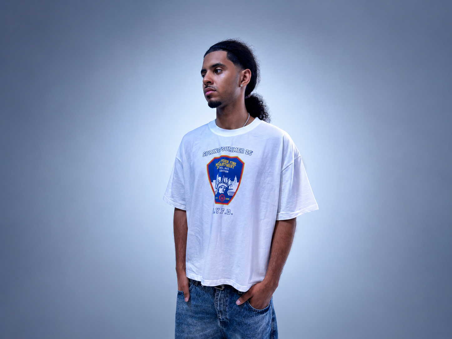 A man wearing a white summit no.26 graphic t-shirt with a blue and red shield emblem on the front, paired with blue jeans, standing against a gray studio background.