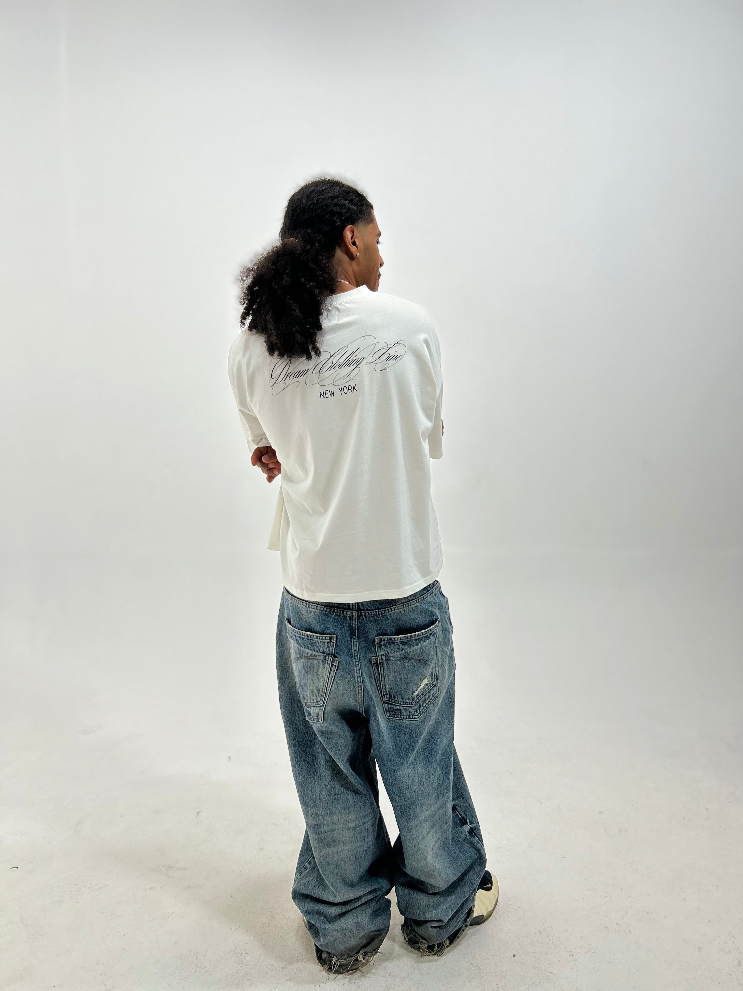A person shown from the back wearing a white oversized T-shirt with black script text on the back and loose blue jeans, standing against a plain light gray studio background.