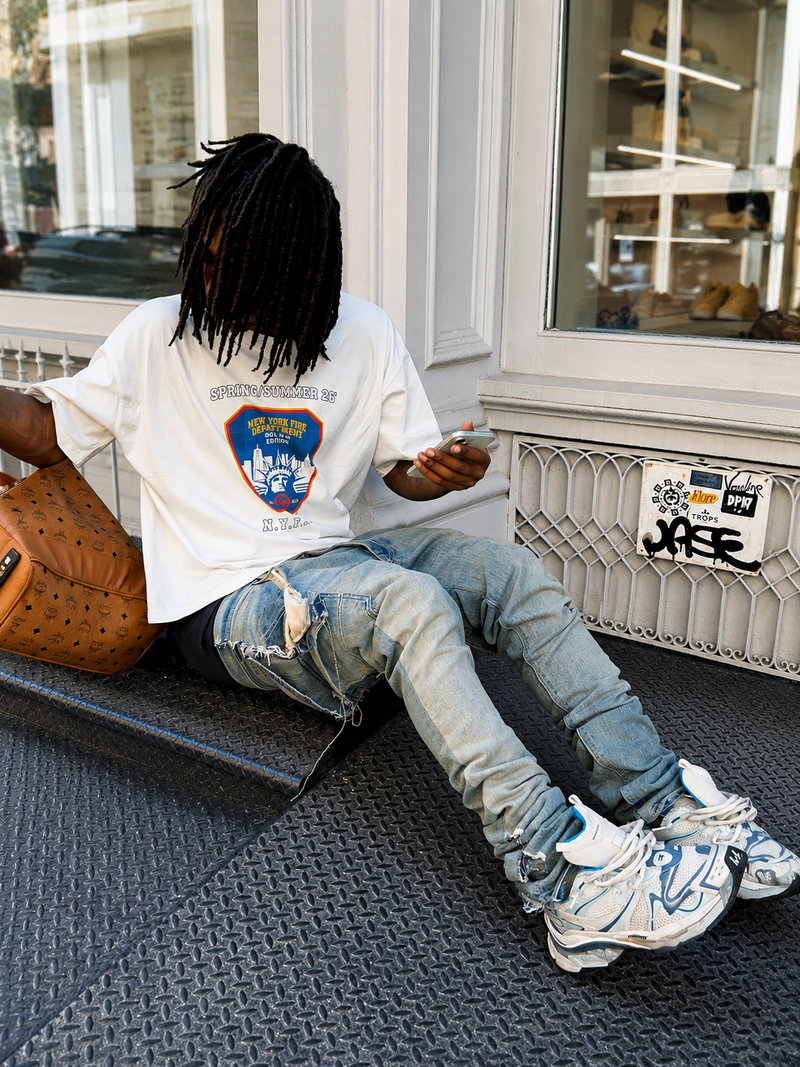 A person sitting on a dark textured step outside a building, wearing an oversized white Summit no.26 graphic T-shirt, ripped light-wash jeans, and white sneakers, with a brown leather bag beside them.