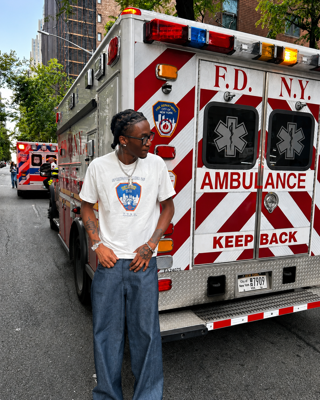 A man wearing a white summit no.26 graphic t-shirt and blue jeans stands in front of the back of an F.D. N.Y. ambulance on a city street.
