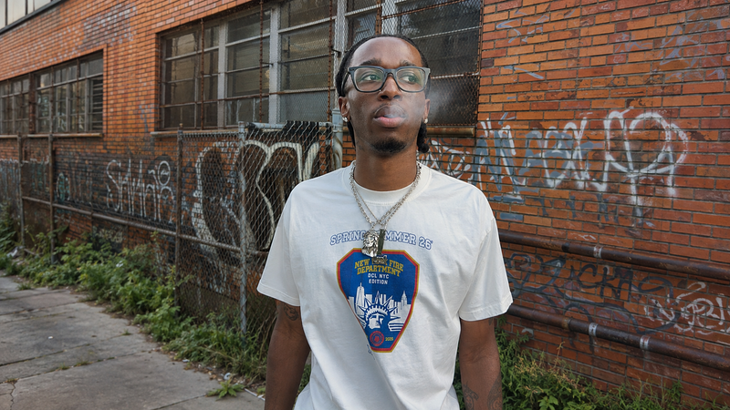 A man wearing a white graphic t-shirt and glasses stands in front of a brick wall covered in graffiti, with smoke visible around his mouth.